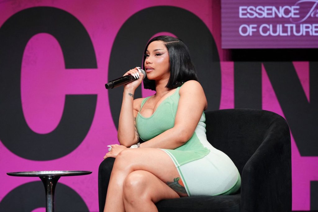 A woman in a light green dress sits on a black chair, speaking into a microphone on stage with a pink and black backdrop that includes the words Essence and Of Culture.