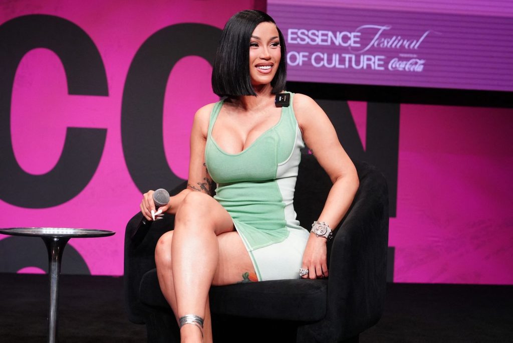 A woman in a green and white dress sits on a black chair, smiling and holding a microphone, with a pink and purple Essence Festival of Culture background behind her.