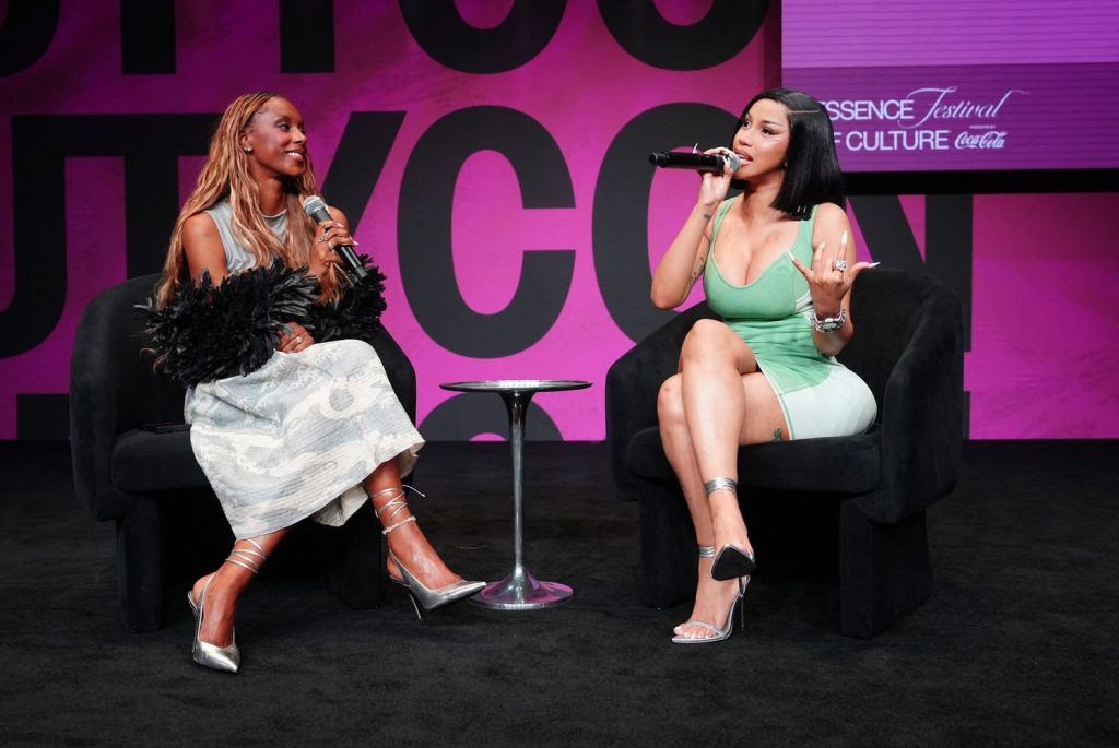 Two women sit on black chairs on stage, speaking into microphones. The background features pink signage with Essence Festival and Coca-Cola logos. A small table sits between them.