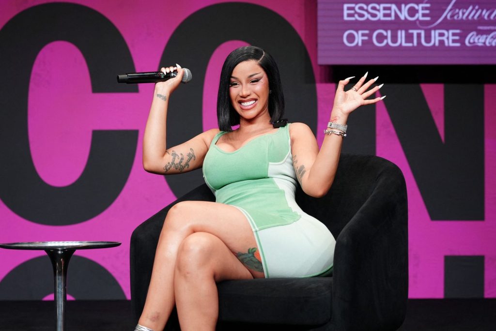 A woman in a green dress sits on a black chair, holding a microphone and smiling with one hand raised. The background is pink with text reading “ESSENCE Festival of Culture.”.