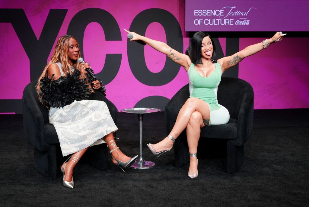 Two women sit on stage at the Essence Festival of Culture. One holds a microphone, while the other smiles, sticking out her tongue and stretching her arms wide. The background is pink with event branding.