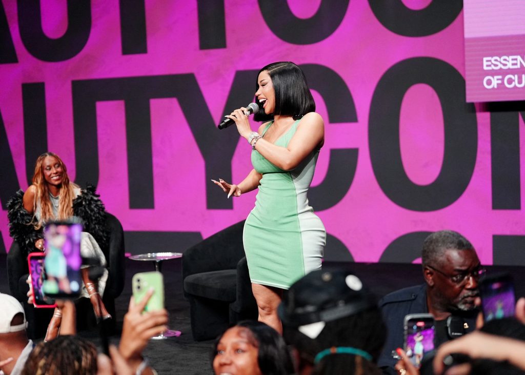A woman in a green and white dress sings on stage with a microphone, while a seated woman smiles nearby. Audience members hold up their phones to take photos. The background features large, bold pink and black text.