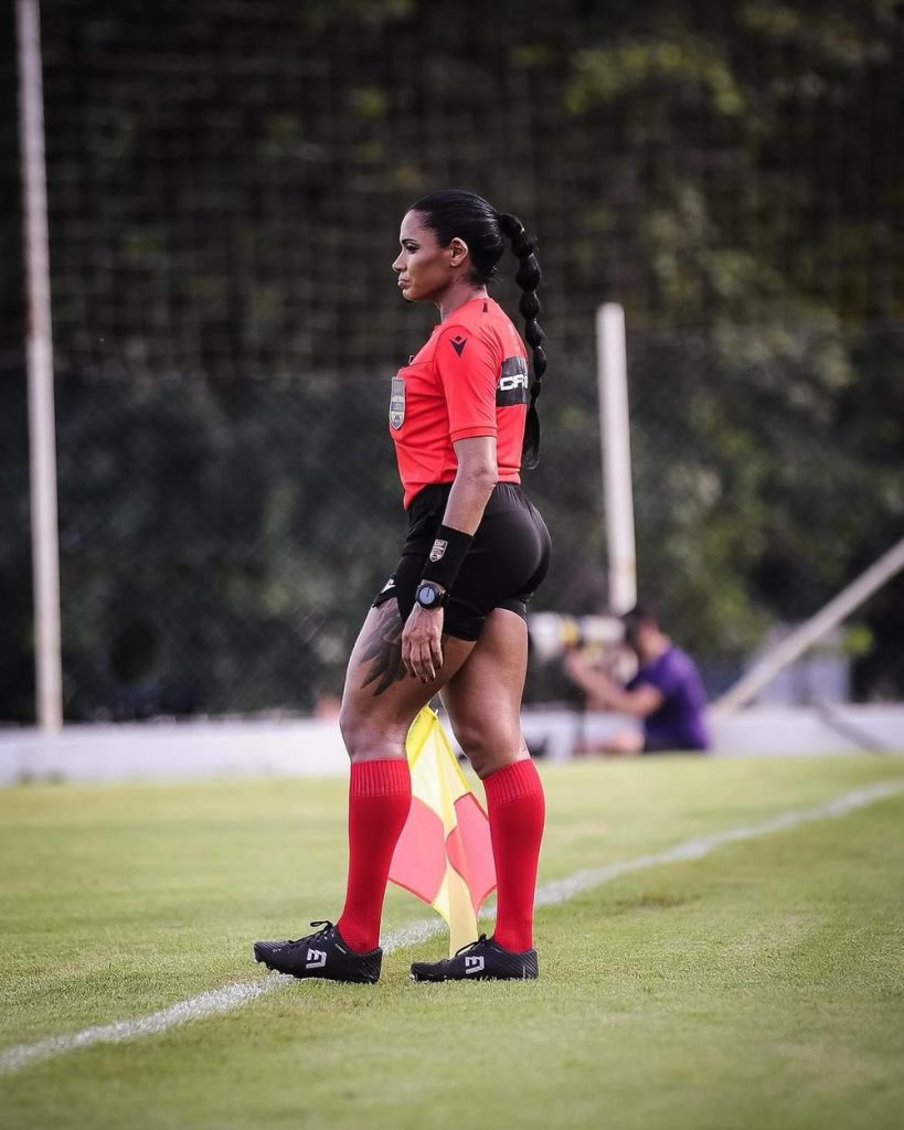 A soccer assistant referee in a red shirt and black shorts stands on the sideline holding a flag, focused on the field during a match.