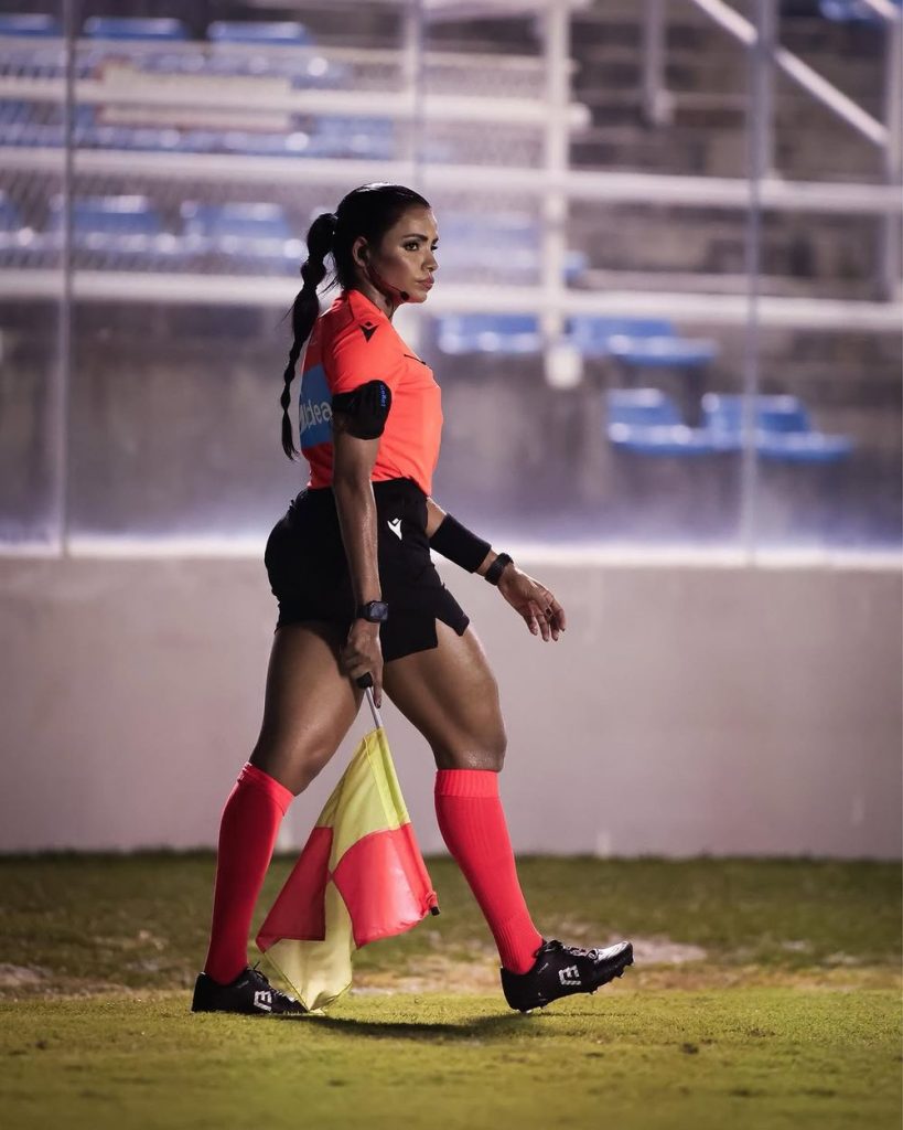 A soccer assistant referee in a red shirt, black shorts, and red socks walks on the field holding a yellow and orange flag. Empty blue stadium seats are visible in the background.