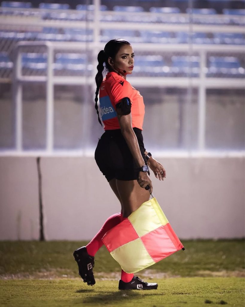 A soccer assistant referee in a red shirt and black shorts holds a checkered yellow and orange flag while walking on the field, looking over her shoulder.