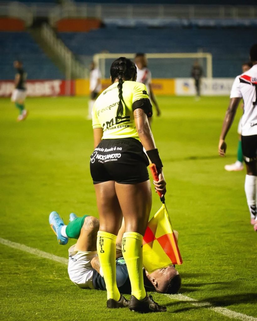 A soccer assistant referee stands near a player lying on the field in front of her, holding a flag. Other players are visible in the background on the field under stadium lights.