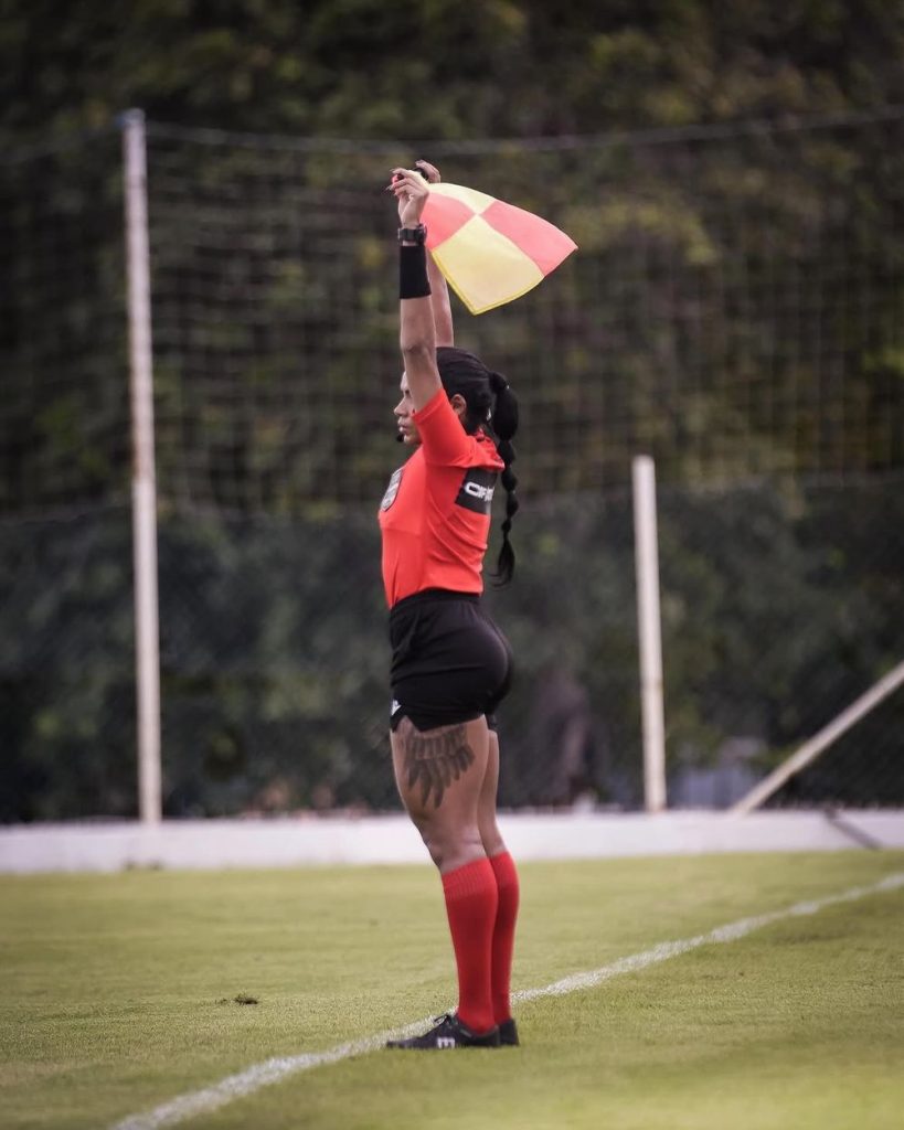 A soccer assistant referee in red raises a checkered flag straight up while standing on the sideline during a match, with a netted fence and greenery in the background.