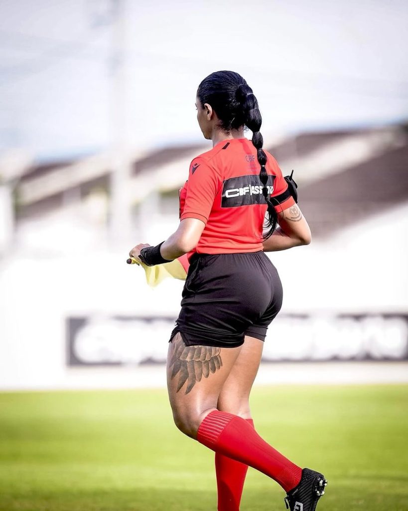 A soccer referee with a long braid, wearing a red shirt, black shorts, and red socks, walks on a grassy field. A large feather tattoo is visible on her thigh. The background is blurred.