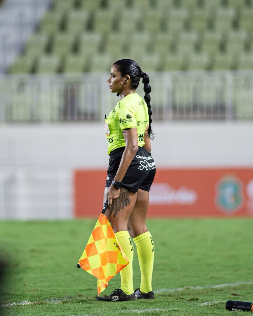 A soccer assistant referee stands on the field holding a checkered yellow and orange flag, wearing a yellow uniform, with a stadium seating area blurred in the background.