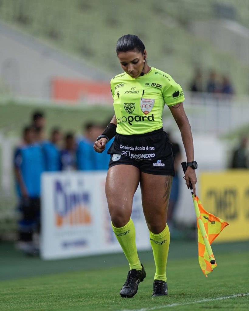 A female soccer assistant referee in a neon yellow uniform walks on the field holding a checkered flag, with players and a blurred crowd in the background.