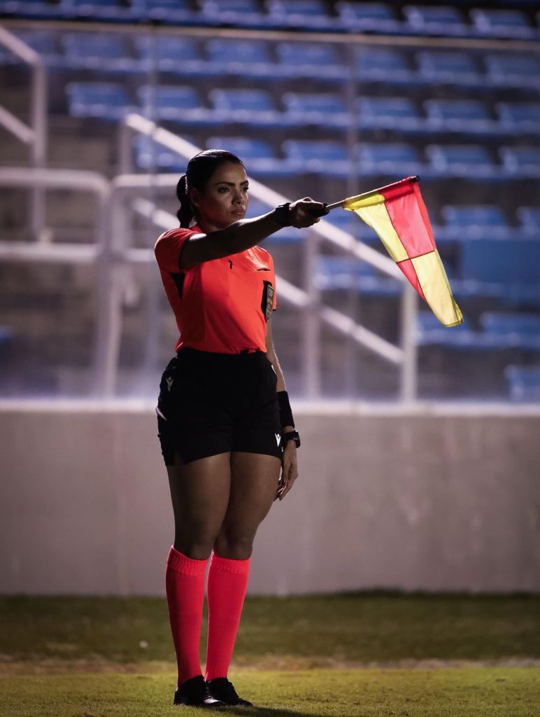 A soccer assistant referee stands on the sideline holding a raised yellow and red flag, wearing a bright orange uniform with black shorts and pink socks, with empty blue stadium seats in the background.