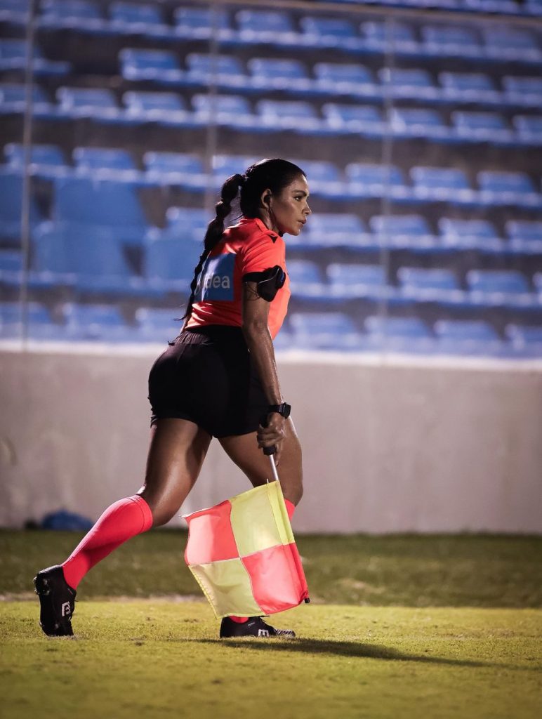 A female soccer assistant referee in a pink uniform holds a yellow and orange flag on a field, with empty blue stadium seats in the background.
