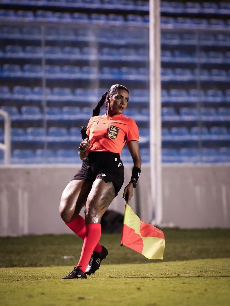 A soccer assistant referee in an orange shirt and black shorts holds a flag while running on the field, with empty blue stadium seats in the background.