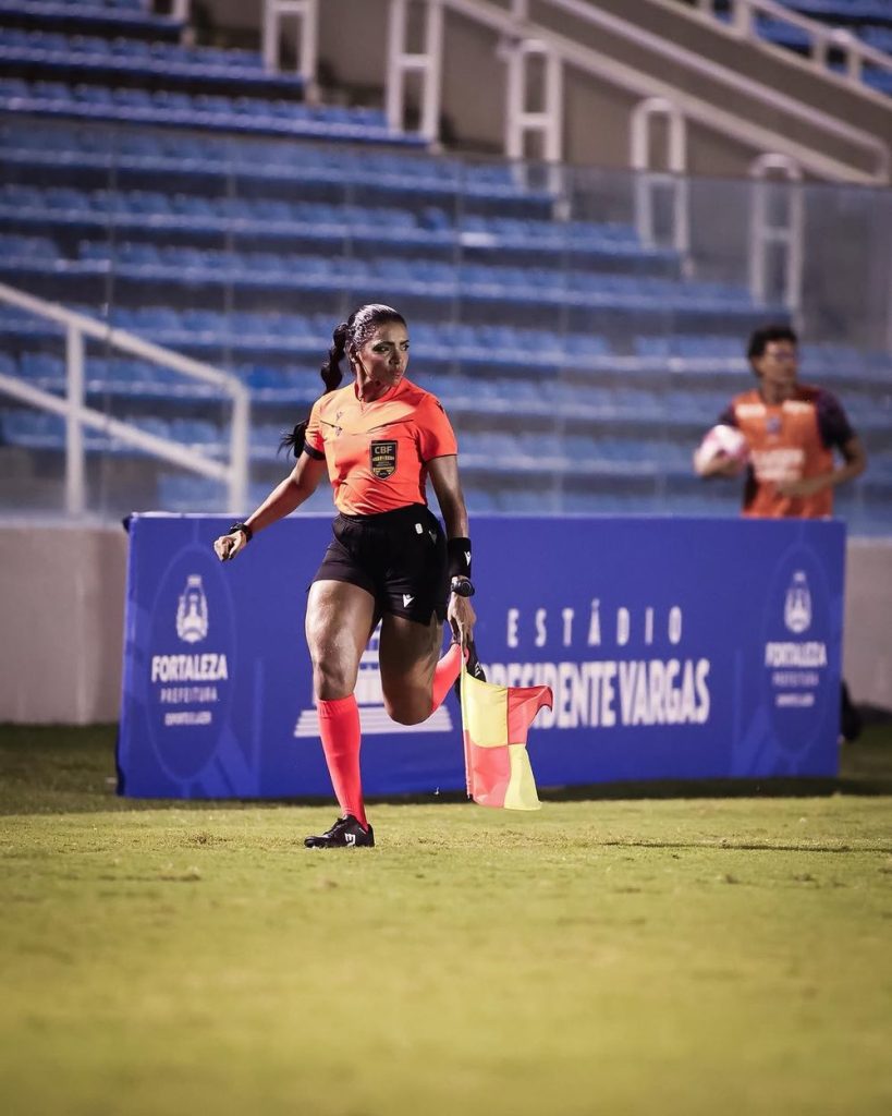 A female soccer assistant referee in a red uniform runs along the sideline holding a flag during a match at a stadium with blue seats.