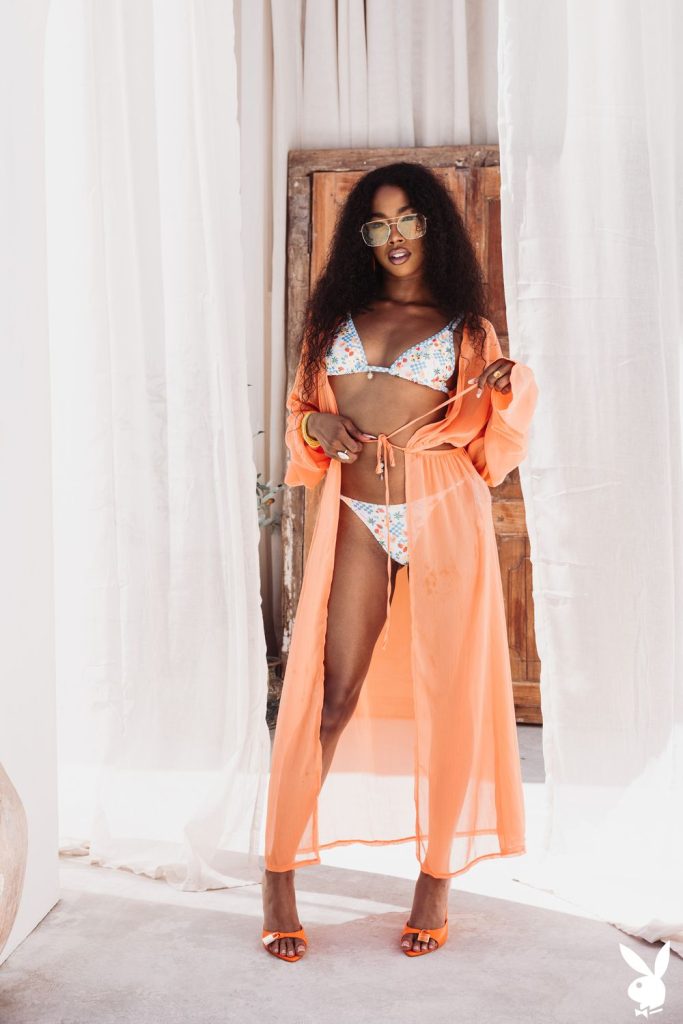 Black woman wearing sunglasses, a floral bikini, and an orange sheer cover-up stands confidently indoors with white curtains and a wooden door behind her.