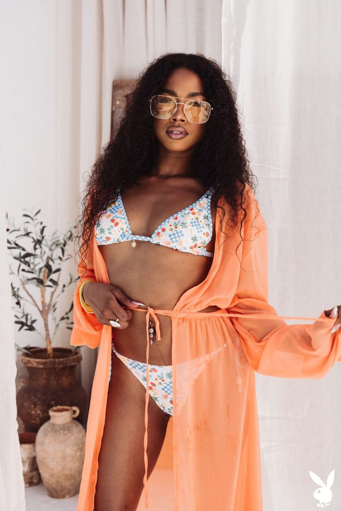 A black woman in sunglasses and a floral bikini with an orange sheer cover-up stands indoors near pottery and a plant, posing confidently.
