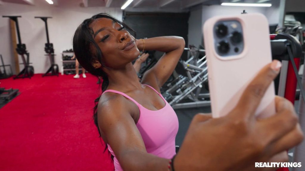 A black woman in a pink tank top takes a selfie at the gym, smiling with one arm behind her head. Exercise equipment and a red carpet are visible in the background.