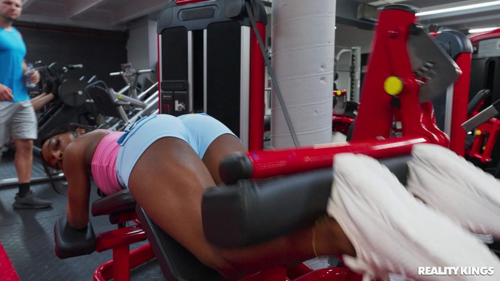 From behind view of a black woman in workout shorts lying face down on a workout bench at the gym. She looks back and exercise equipment and a man is walking by in the background.