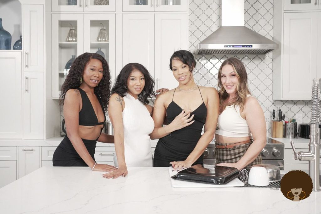 Four women pose and smile together in a modern kitchen, standing behind a white counter with a laptop and mug visible in the foreground.