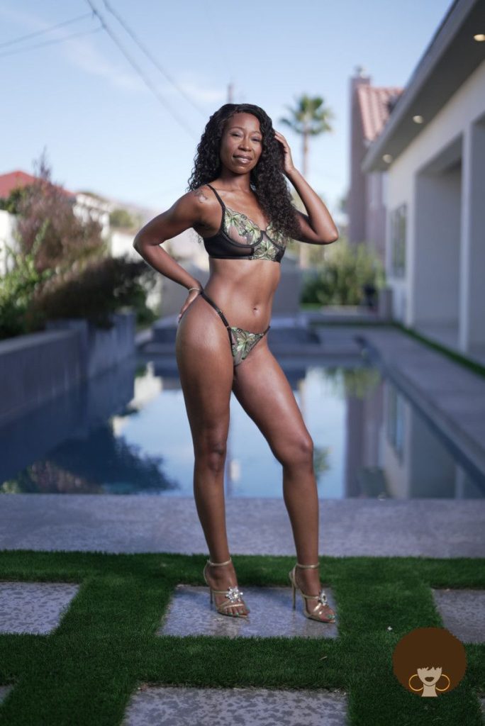 A woman in a green bikini and heels stands confidently by a backyard pool, with houses, plants, and palm trees visible in the background on a sunny day.