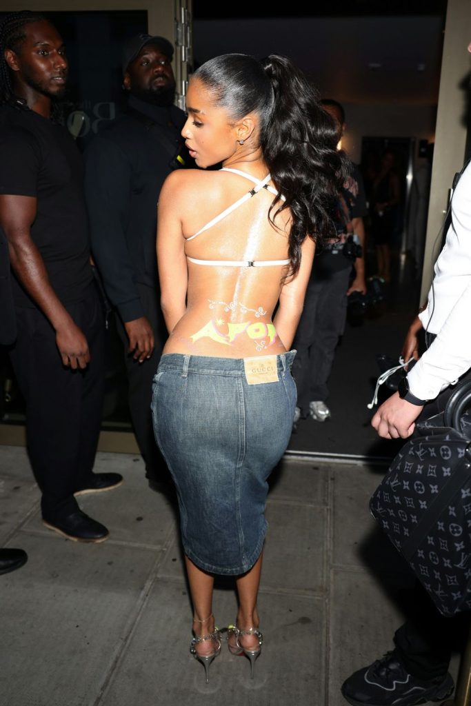 A woman with a high ponytail wears a backless, strappy top, exposing a large back tattoo, and a knee-length denim skirt with heels, standing outside at night among several people.