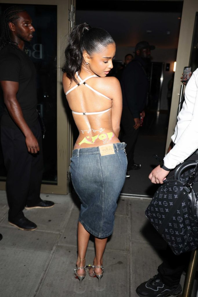A woman in a backless top and denim skirt stands outside, looking over her shoulder. Several people are nearby, and she appears to be entering or exiting a building at night.
