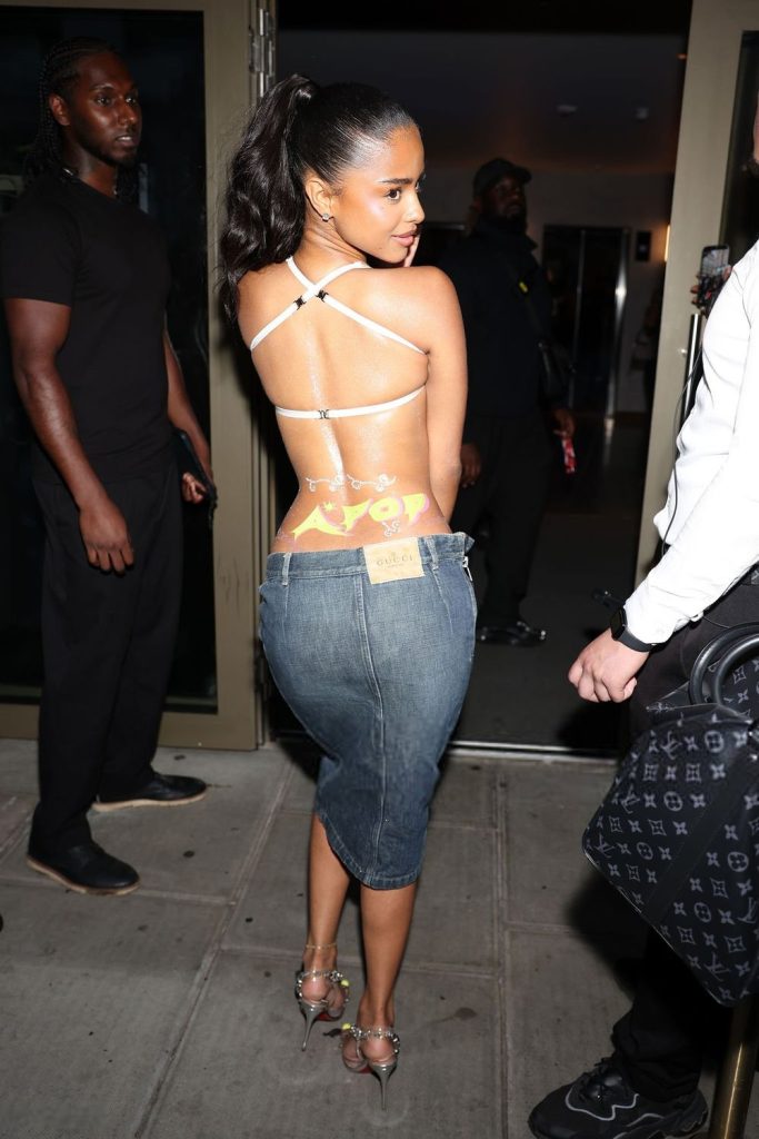 A woman poses outside, wearing a jeweled strappy back top and a knee-length denim skirt, with her hair in a high ponytail. People stand nearby as she looks over her shoulder, smiling at the camera.