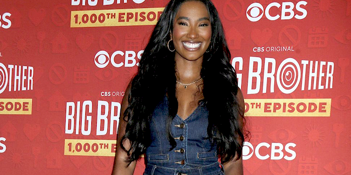 Taylor Hale in a denim dress smiles in front of a red Big Brother 1,000th Episode CBS backdrop.
