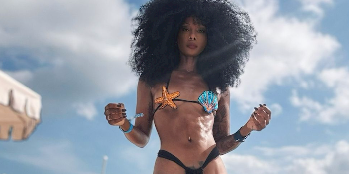 A woman with curly hair poses on a sandy beach, wearing a bikini with starfish and seashell designs. Lounge chairs and umbrellas line the background under a partly cloudy sky.