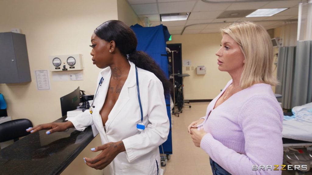 A Black female doctor wearing a white lab coat with a stethoscope and a blonde woman in a light purple sweater stand at a counter in a hospital room.