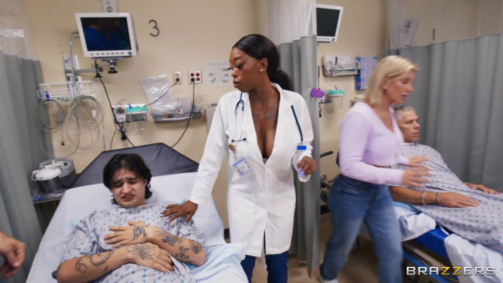 In a hospital room, a female doctor in a white coat stands between two patients in hospital gowns. To the right, a blonde woman wearing a light pink top and blue jeans leans toward the patient in the second bed.