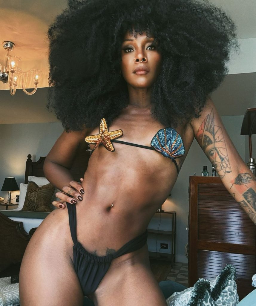 A woman with voluminous curly hair poses indoors wearing a bikini top decorated with a starfish and a seashell, and black bikini bottoms. She stands confidently next to a bed in a stylish room.