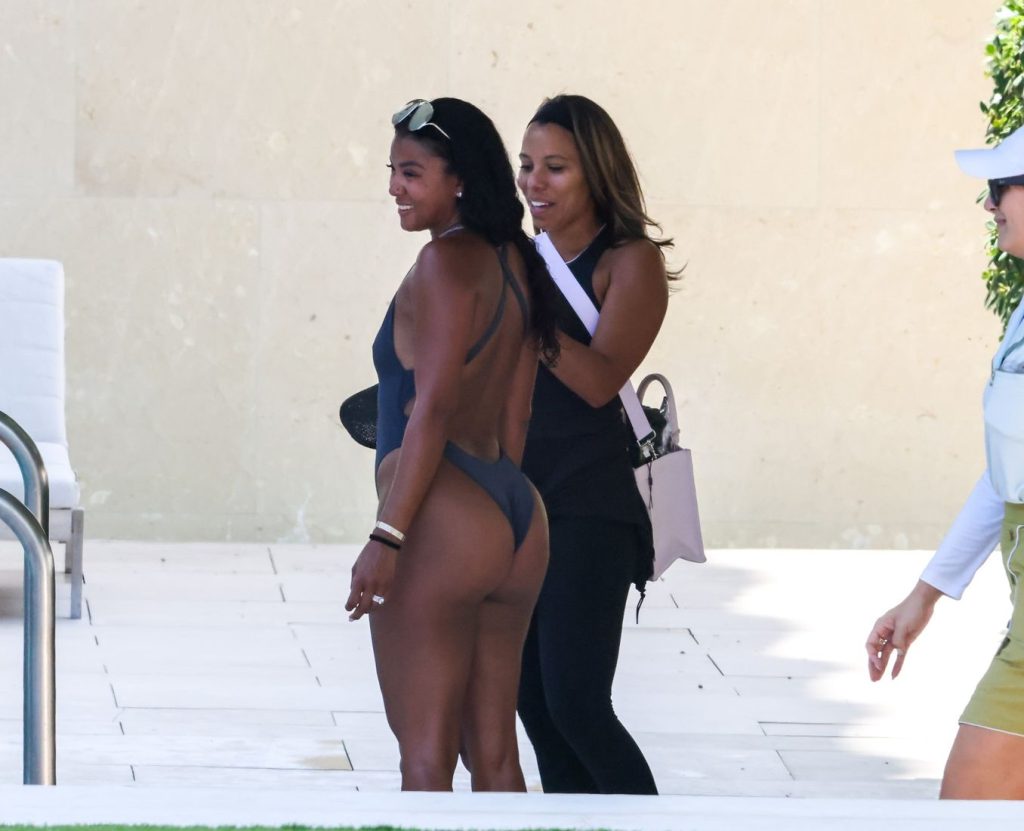 Two women stand by a poolside. One wears a dark one-piece swimsuit and holds a hat, the other wears black athletic clothing and carries a light bag. Both are smiling and talking.