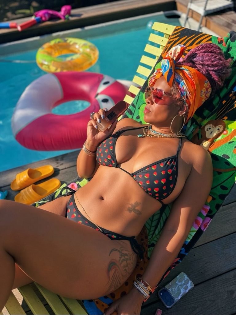 A woman in a black bikini with red hearts lounges on a chair by a pool, holding a popsicle. She wears sunglasses, a colorful headscarf, and jewelry. Pool floats and sandals are nearby.