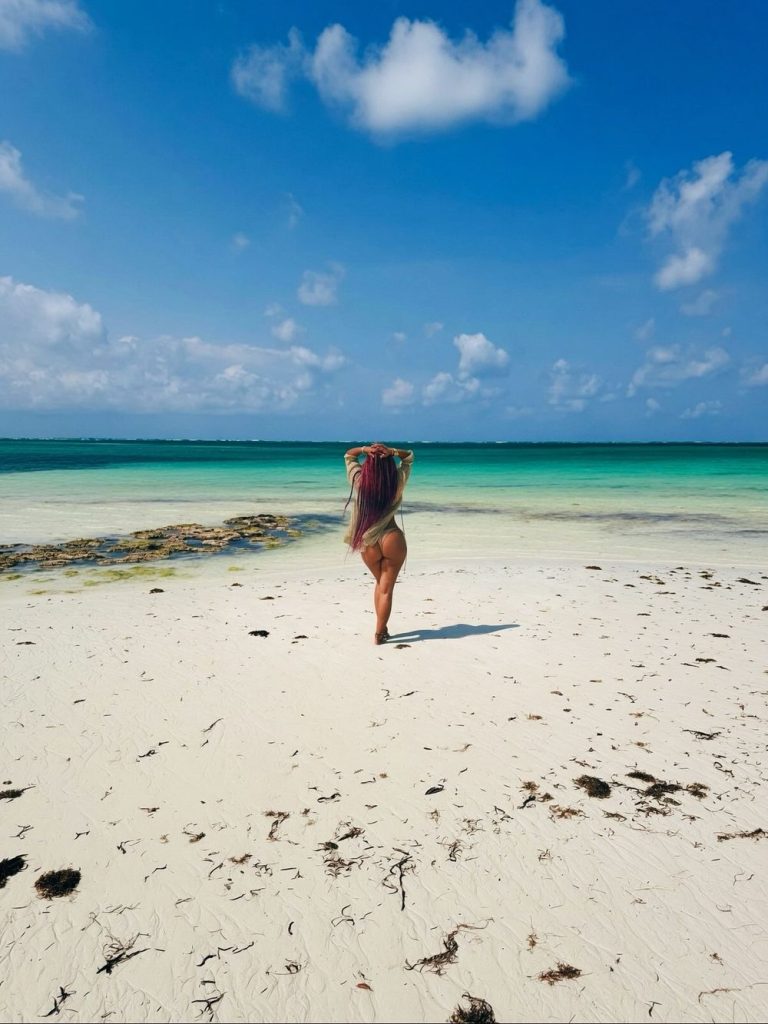 A person with long hair stands on a white sandy beach facing the turquoise sea, with blue sky and clouds above. Seaweed is scattered on the shore.