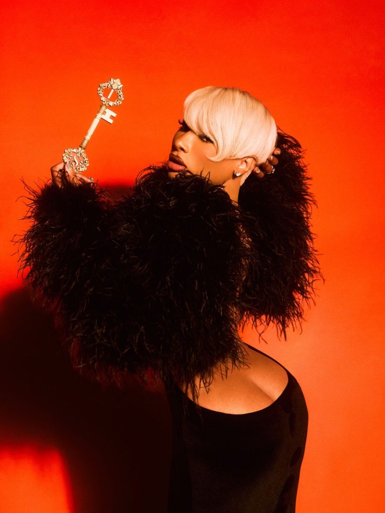 A woman with platinum blonde hair and dramatic black feathered sleeves poses against a bright orange background, holding a large ornate key over her shoulder.