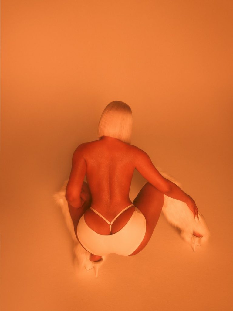 A woman with short blonde hair, seen from behind, squats on the floor wearing white high heels and minimal white clothing, draping a white fur garment in an orange-tinted setting.