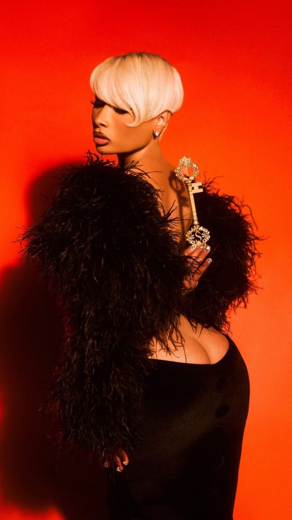 A woman with short platinum blonde hair wears a black feathered jacket and holds a gold key, posing against a vibrant red background.