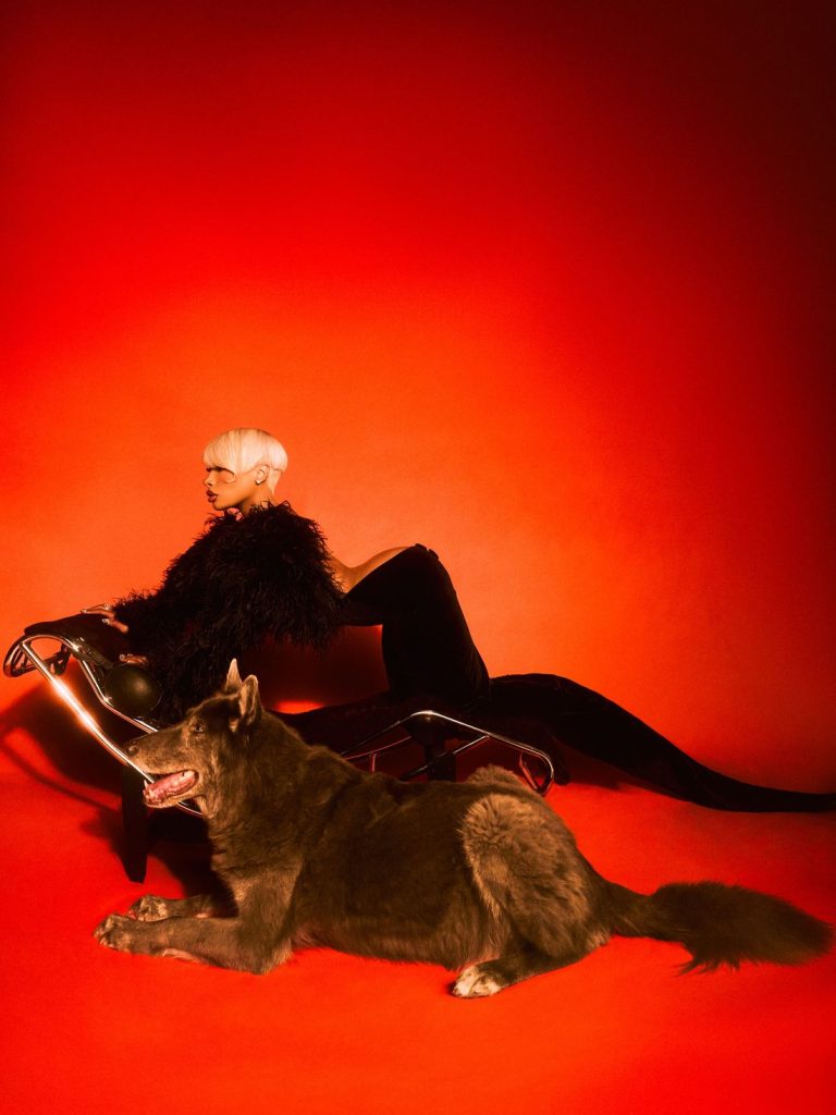 A woman with short platinum hair, dressed in black with feathered sleeves, lounges on a modern chair. A large black dog rests in the foreground. The background is a dramatic, vivid red.