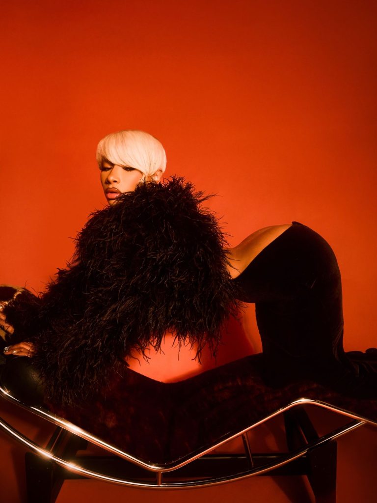 A woman with short blonde hair and a black feathered jacket poses on a modern chaise lounge against a bold red background, creating a dramatic and stylish scene.
