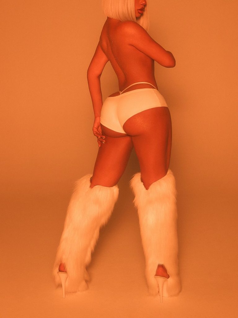 A woman with a short blonde wig poses topless, wearing white underwear and tall white furry boots, against a warm orange background.