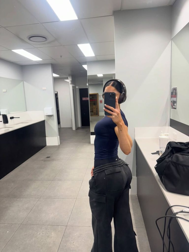 Woman in dark clothing takes a mirror selfie in a modern, well-lit gym bathroom, wearing headphones and holding a phone. Gym bags and a drink cup are visible on the counter nearby.