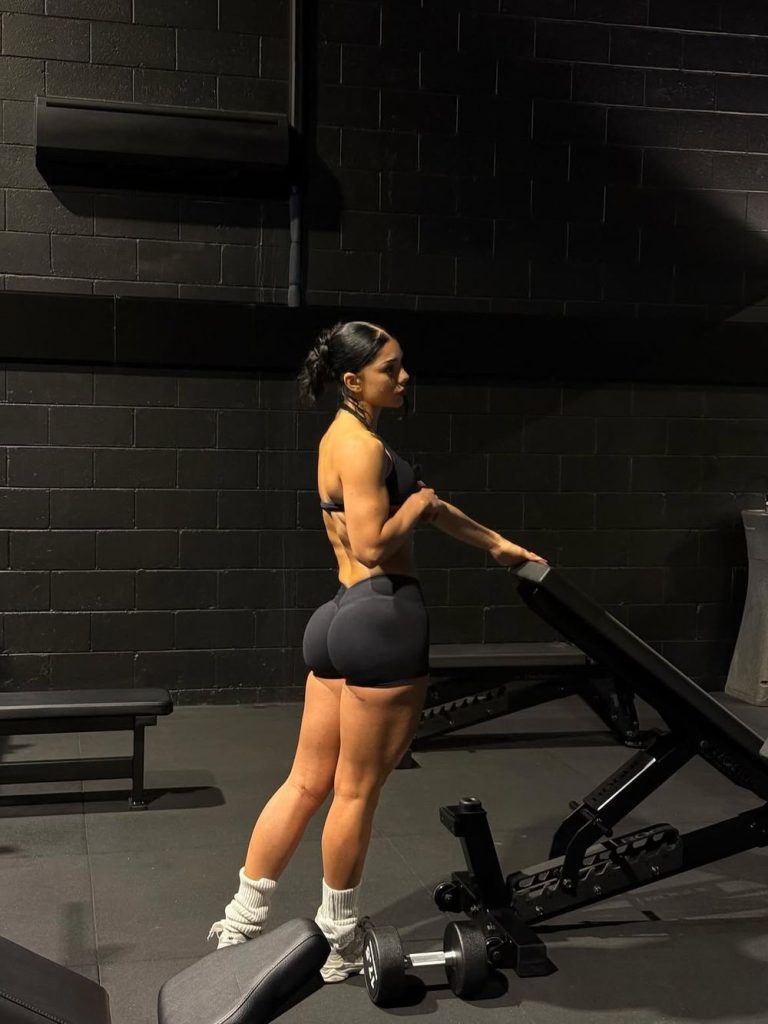 A woman in athletic wear stands beside a bench in a gym, holding its backrest. She faces sideways, showing her muscular build. Gym equipment and a black wall are visible in the background.