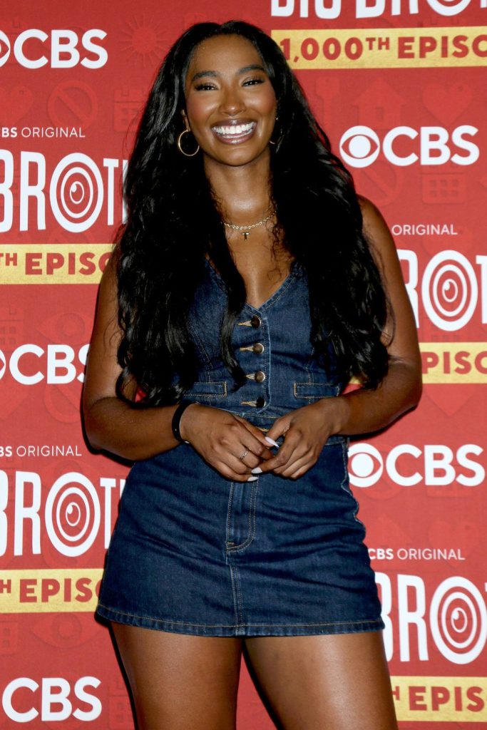 A woman in a sleeveless denim dress smiles in front of a red CBS Big Brother 25th season backdrop.