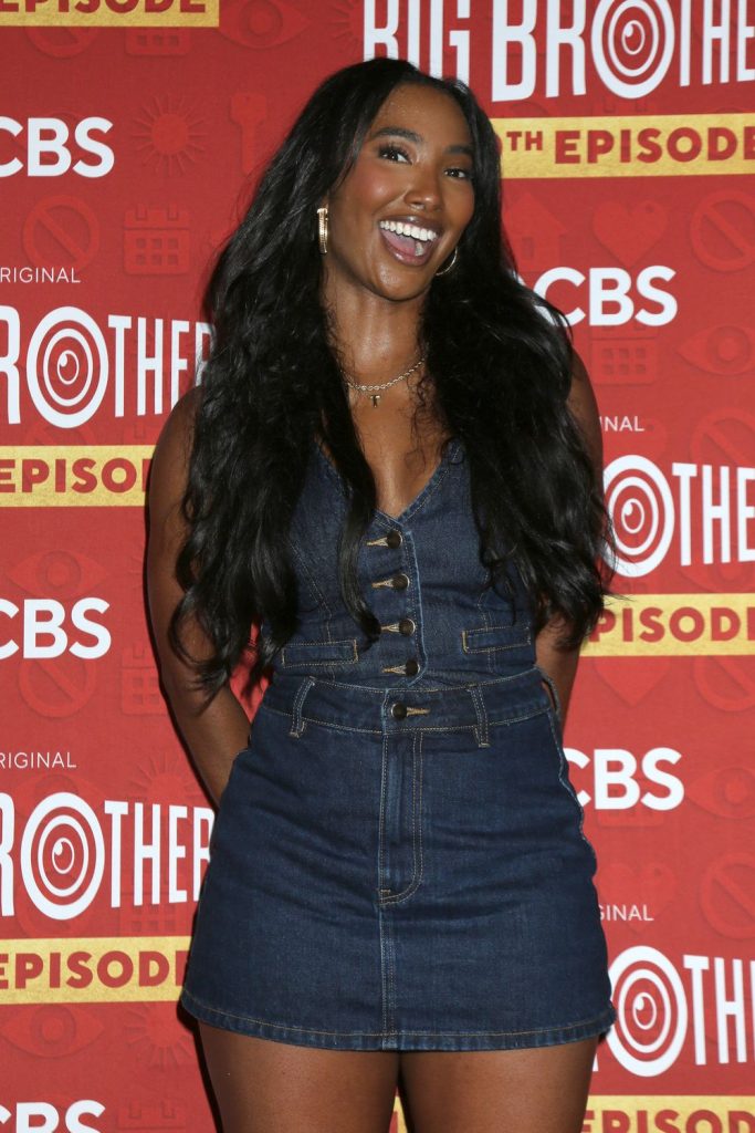 A woman with long black hair in a denim dress smiles in front of a red Big Brother 25th episode backdrop featuring CBS logos and yellow text.