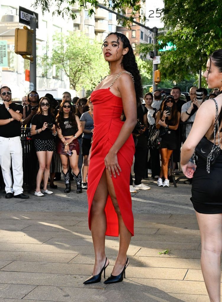 A woman in a red dress with a thigh-high slit and black heels stands confidently on a city street, surrounded by a crowd of onlookers, some taking photos. Trees and buildings are visible in the background.