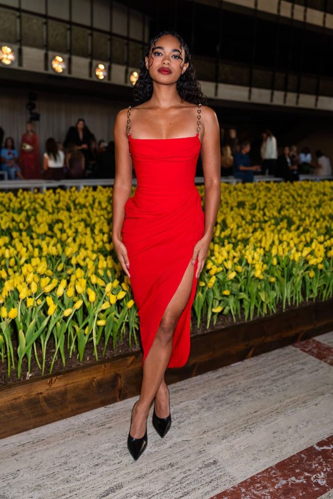 A woman in a red dress with chain straps stands confidently in front of a bed of yellow tulips, indoors, with people seated in the background. She wears black heels and has curly hair.