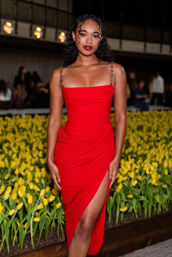 A woman in a red dress with chain straps poses confidently in front of a bed of yellow tulips, with people and lights blurred in the background.