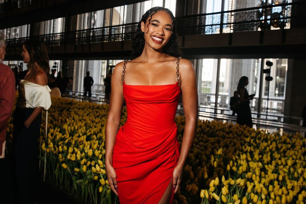 A woman in a red dress stands smiling in front of a large display of yellow tulips indoors. Other people are visible in the background, along with large windows and modern architectural details.