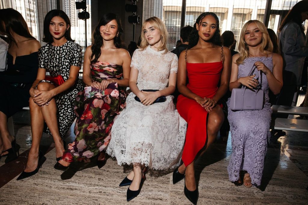 Five women sit in a row at a fashion event, each dressed in elegant, fashionable attire, including floral, lace, red, and lavender dresses, with stylish accessories. They appear poised and are facing the camera.